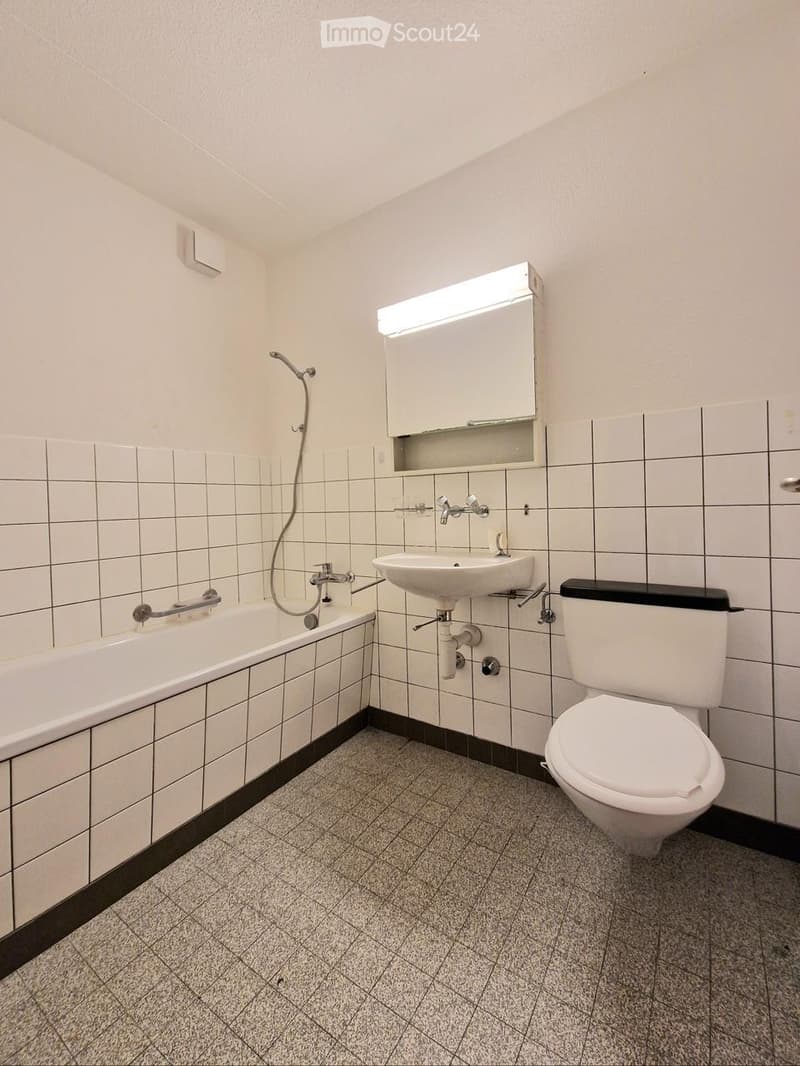 Modern bathroom with white tiled walls, a bathtub, toilet, sink, and a built-in mirror cabinet with lighting above the sink.