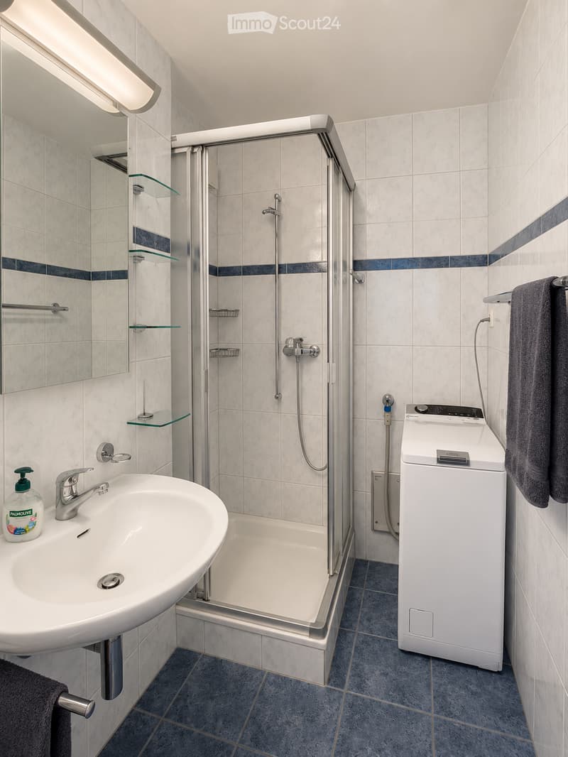 Modern bathroom with a white sink, mirror, glass shelves, a shower with a glass enclosure, and a washing machine. Blue tiles on the floor and walls.