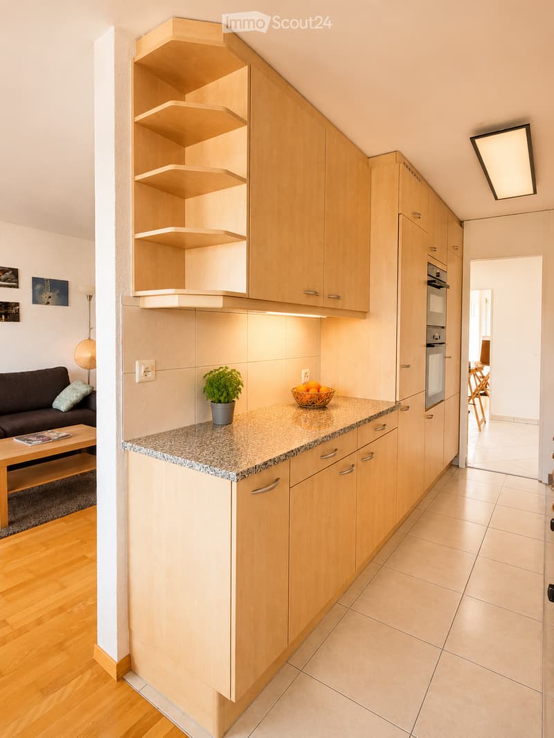 Modern kitchen with light wooden cabinets, granite countertop, and built-in oven. A small living area with a sofa and coffee table is visible to the left.