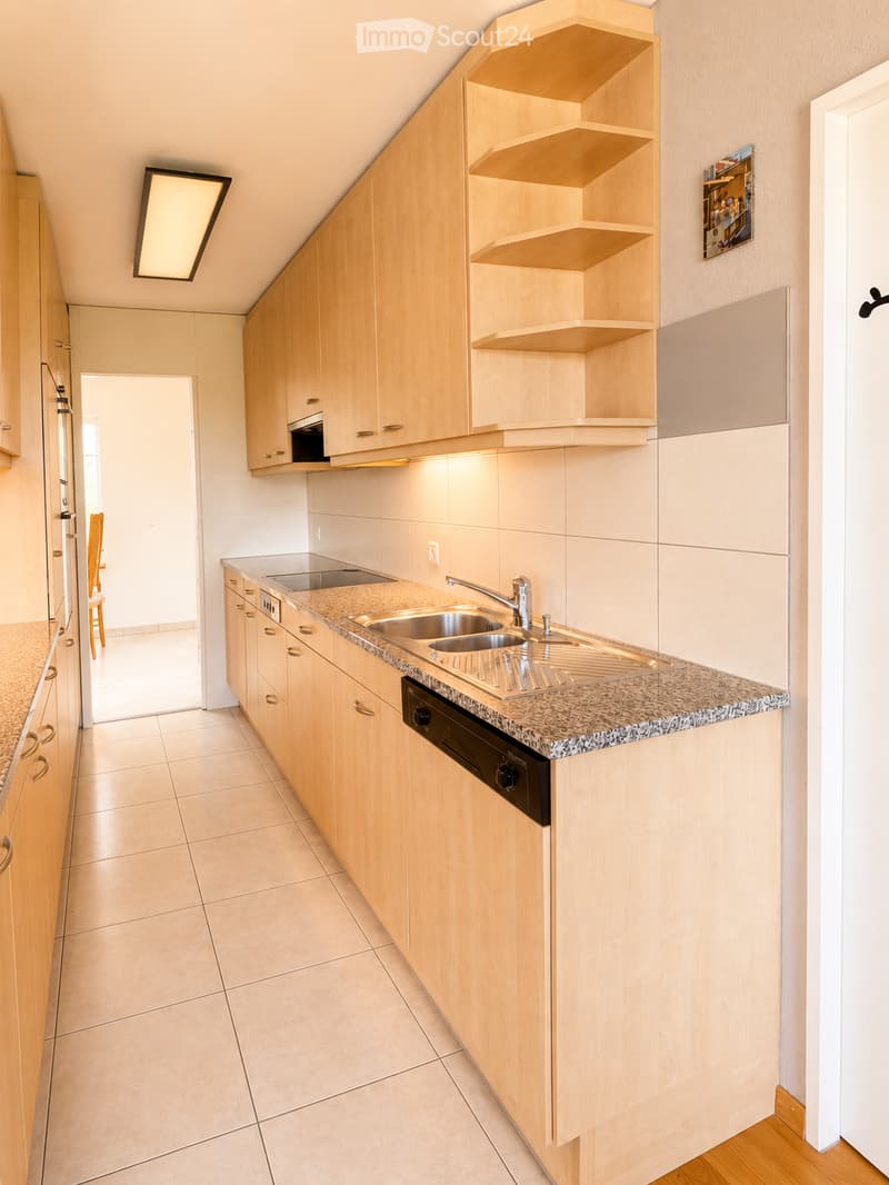 Modern kitchen with light wooden cabinets, granite countertop, stainless steel sink, and built-in oven. Shelves and a small dining area visible in the background.