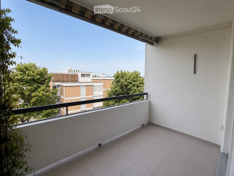Balcony with tiled floor, white walls, black railing, and a view of surrounding buildings and greenery.