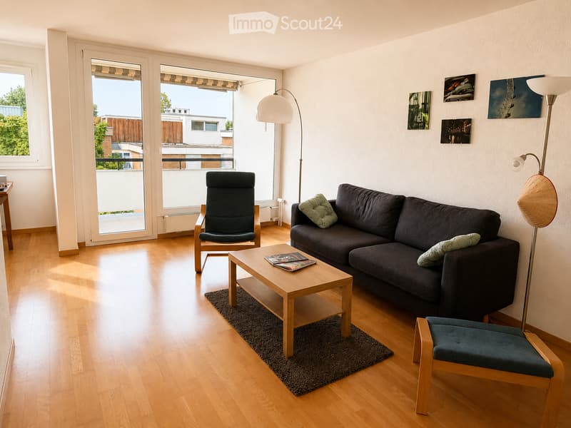Bright living room with wooden flooring, large windows, and a balcony. Features a dark grey sofa, wooden coffee table, two chairs, and a floor lamp.