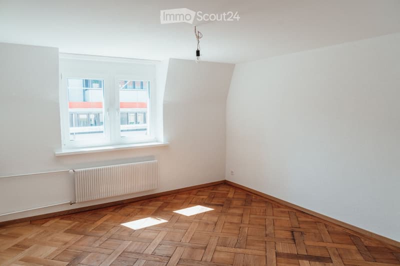 Empty room with wooden herringbone flooring, white walls, large window with white frame, radiator below window, and a single light fixture hanging from the ceiling.