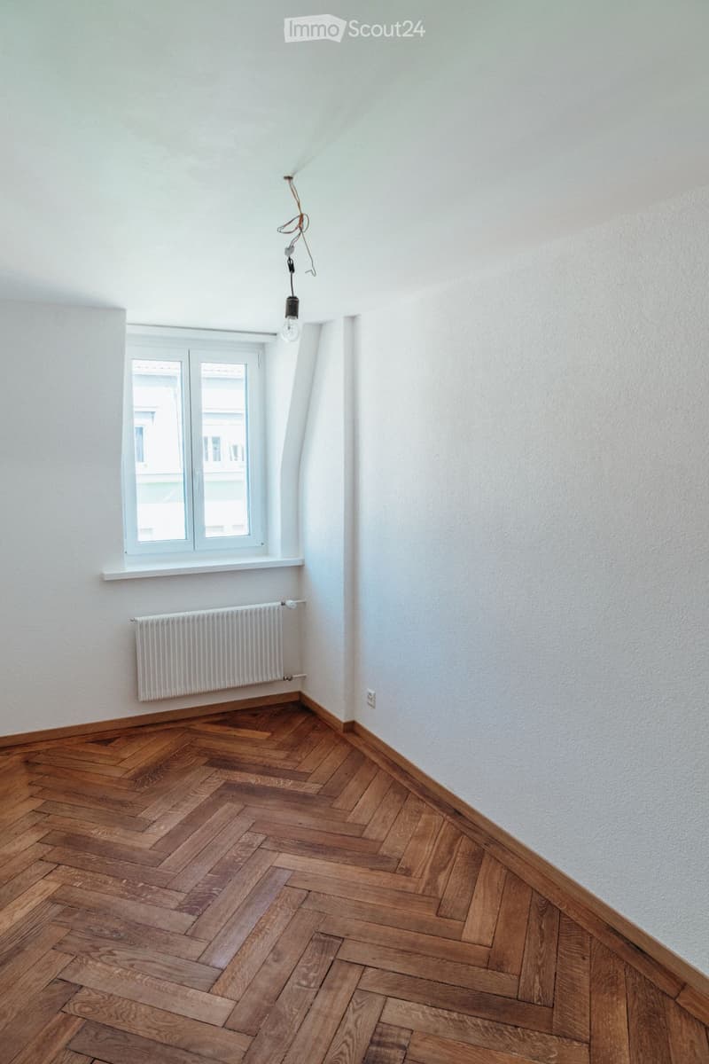 Empty room with wooden herringbone flooring, white walls, a window with white frame, a radiator, and a single light fixture with a bulb hanging from the ceiling.