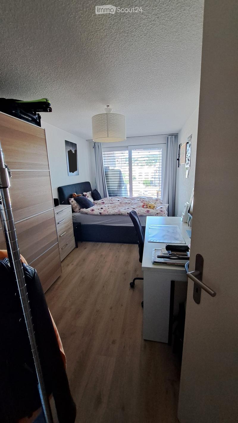 Cozy bedroom with a bed, nightstand, desk, and chair. Features wooden flooring, a large window with blinds, and a hanging lamp.