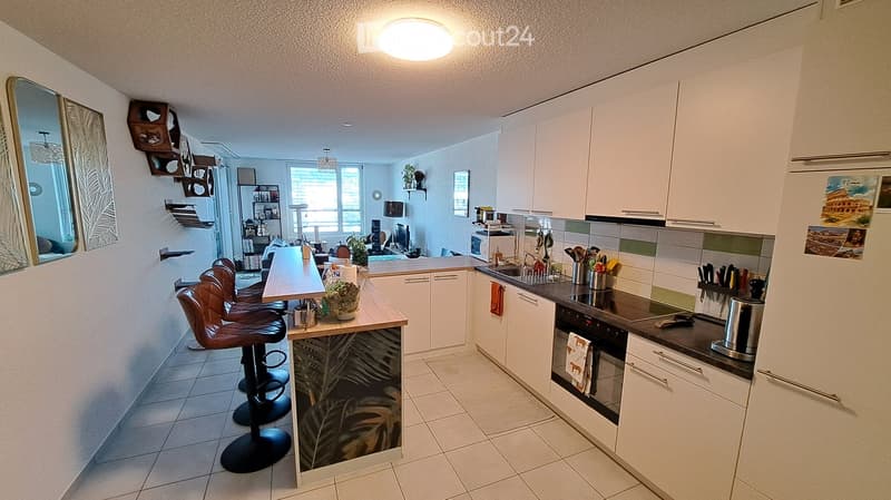 Modern kitchen with white cabinets, a central island with bar stools, and various appliances including an oven, sink, and refrigerator. The space features a tiled floor and large windows, with decorative elements such as a mirror, picture frames, and a sm