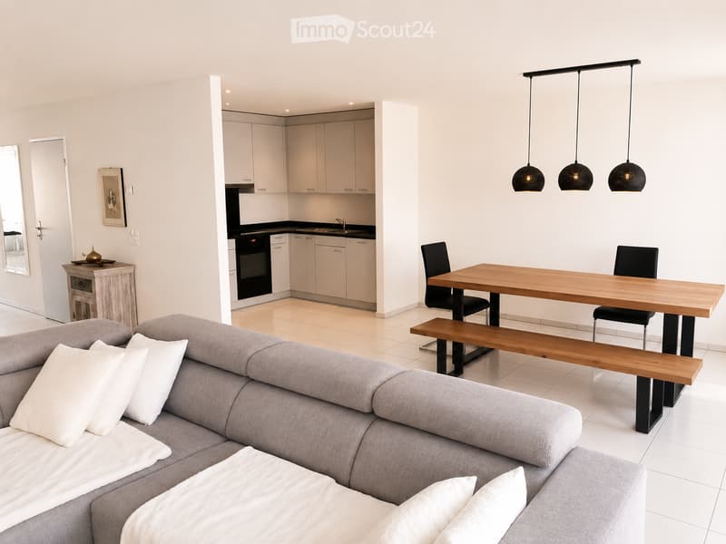 Modern living and dining area with a grey sectional sofa, wooden dining table with bench and chairs, kitchen area with cabinets and appliances, and pendant lights.