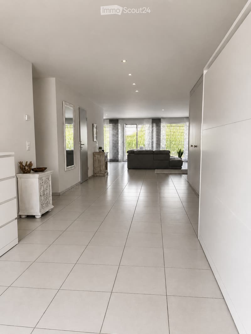 Bright, modern hallway with tiled flooring, white walls, recessed lighting, and a view into a living room area with a sofa and large windows.