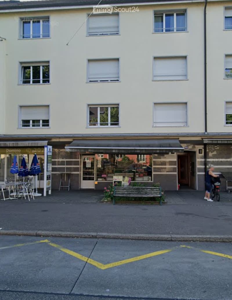 Two-story building with light-colored facade, multiple windows, and a storefront with tables and chairs outside.
