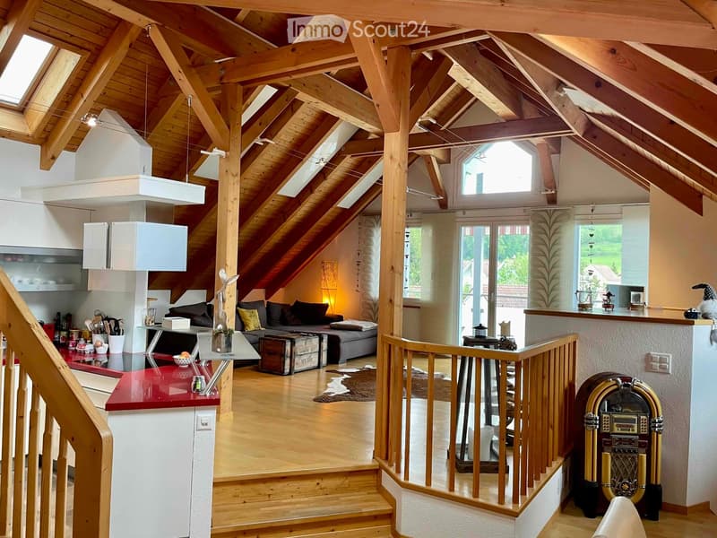 Open-plan attic space with exposed wooden beams, high ceilings, and skylights. Features a living area with a sofa, coffee table, and decorative items, a kitchen area with a red countertop, and a staircase leading to another level.