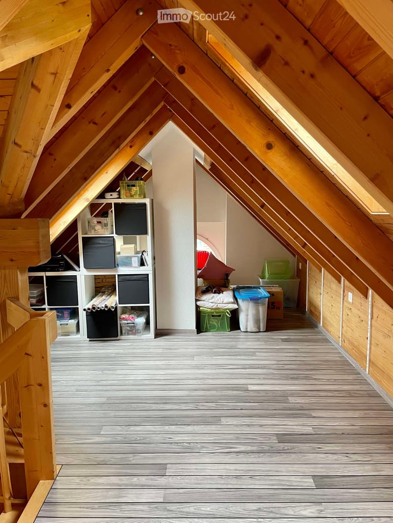 Attic room with wooden beams and slanted ceilings, featuring a wooden floor, a white shelving unit with storage boxes, and a small seating area with a pillow and trash bin.