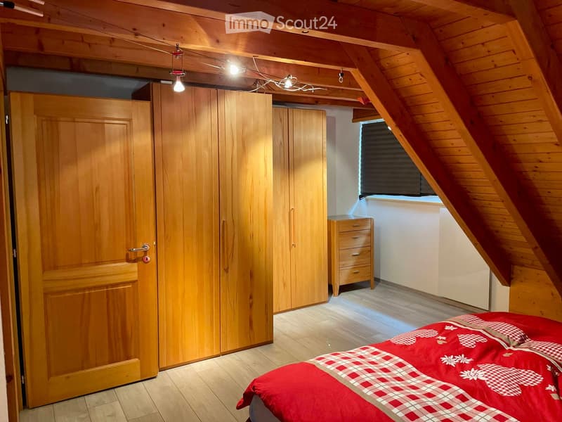 Cozy attic bedroom with wooden walls and ceiling, featuring a large bed with a red and white checkered duvet, a wooden wardrobe, and a small wooden drawer unit near the window.