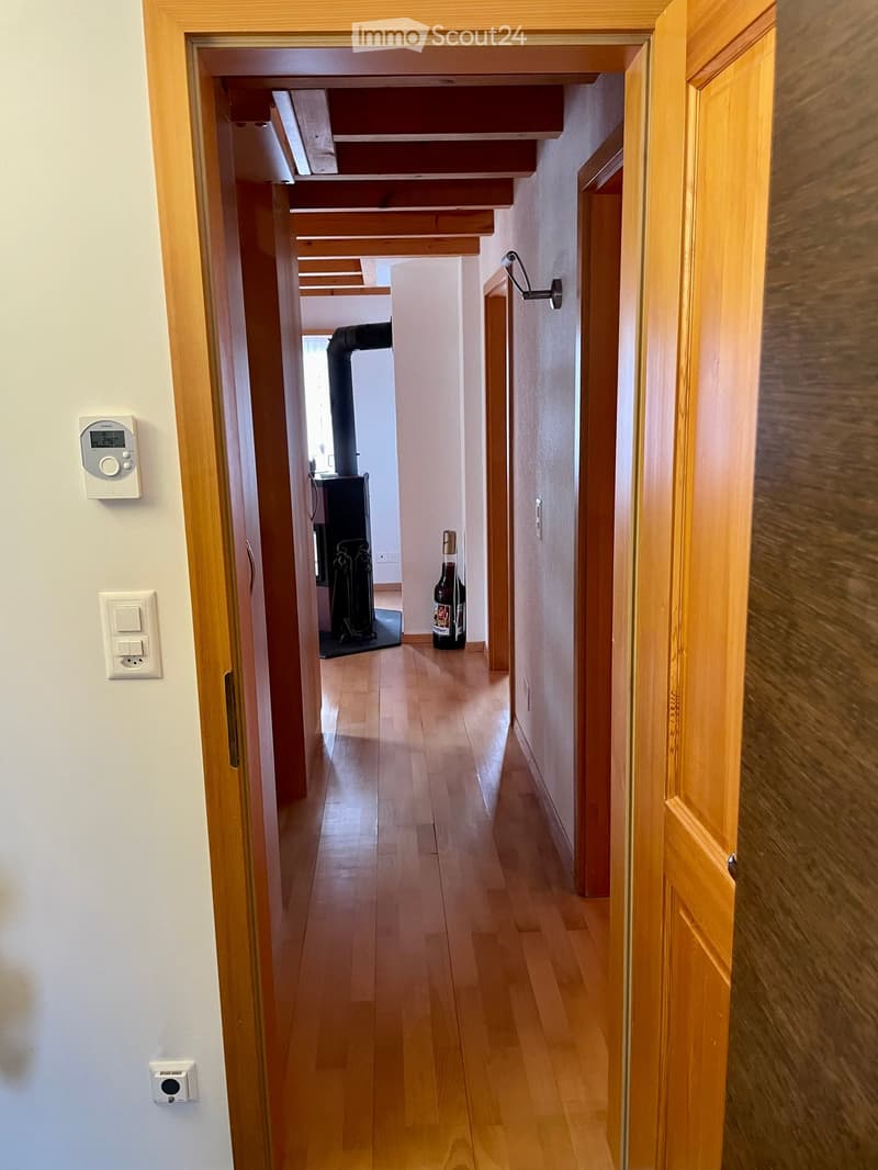 A narrow hallway with wooden flooring and white walls, featuring a thermostat and light switches on the left wall, and a partially visible room with a fireplace and a bottle on the floor in the background.