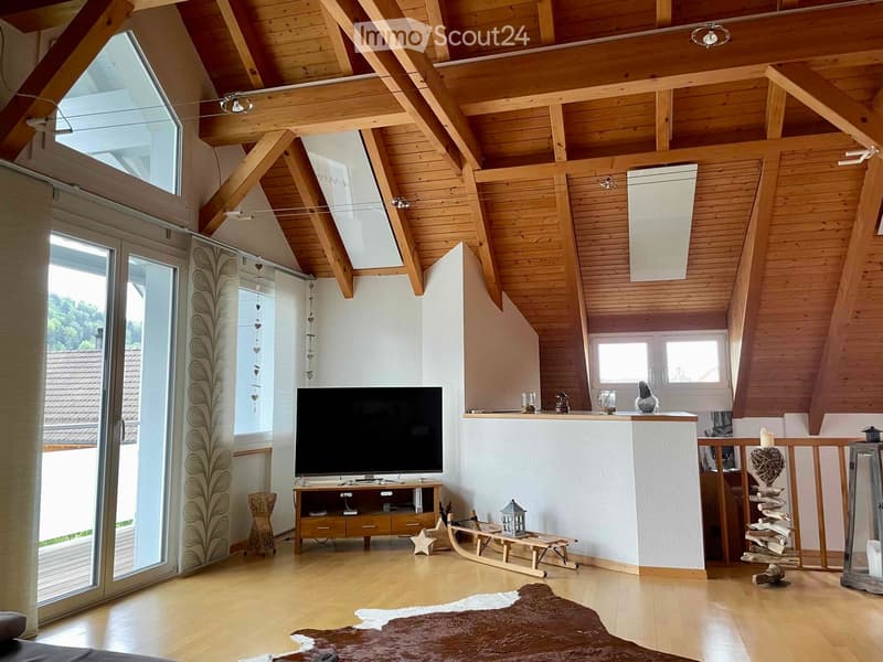 Bright living room with wooden beams and slanted ceiling, featuring a television on a wooden stand, a small wooden chair, a decorative sleigh, and a fur rug on the floor.