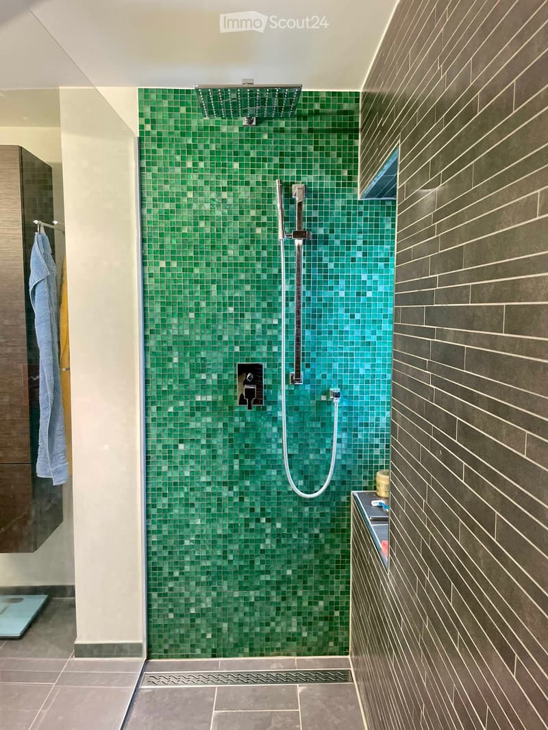 Modern bathroom with green mosaic tiles in the shower area, dark grey tiles on the walls, a towel hanging on a rack, and a small cabinet with toiletries.
