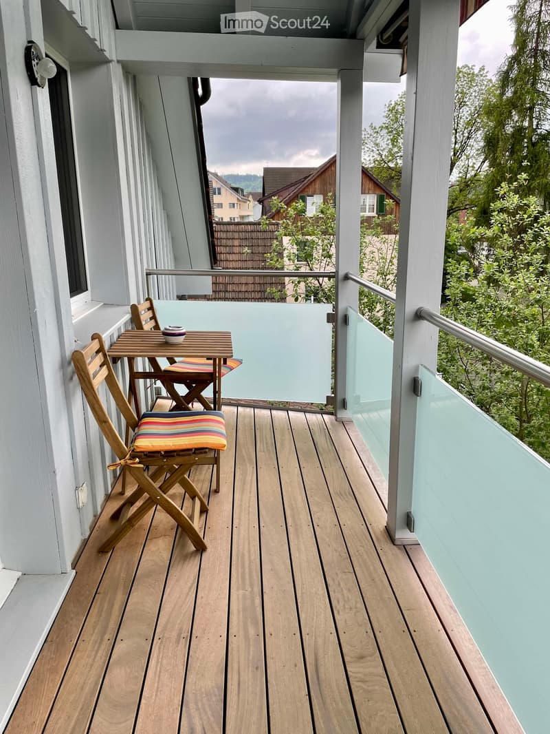 Balcony with wooden decking, glass railing, small table with two chairs, and a cup on the table.