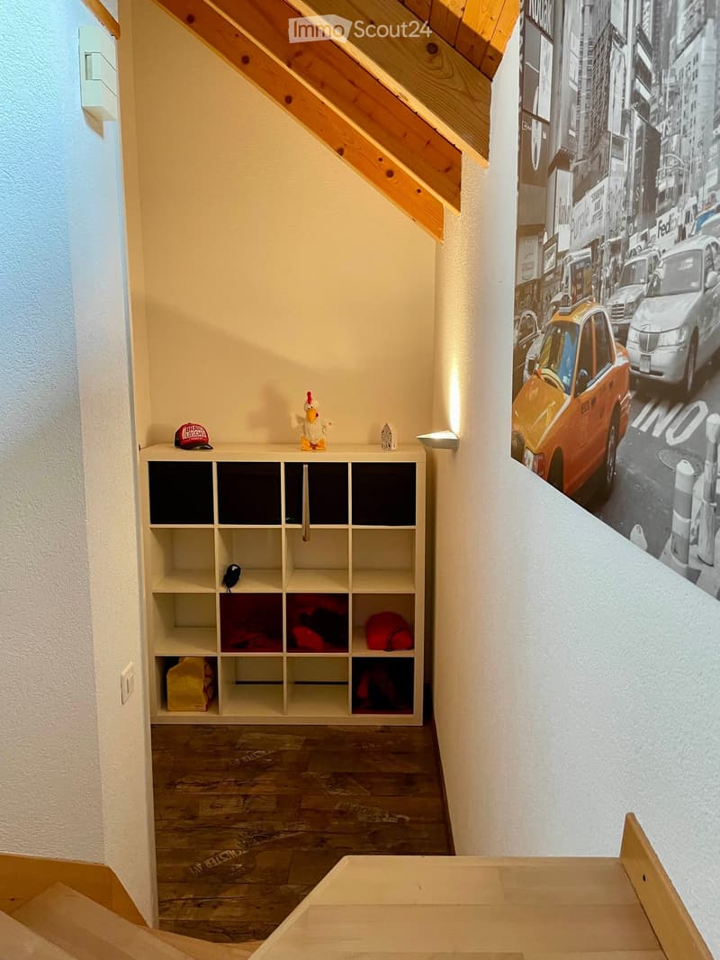 A narrow hallway with wooden flooring, white walls, and a large mural of a city street scene. A white shelving unit with multiple compartments is visible, containing various items such as hats, bags, and a small decorative figure.