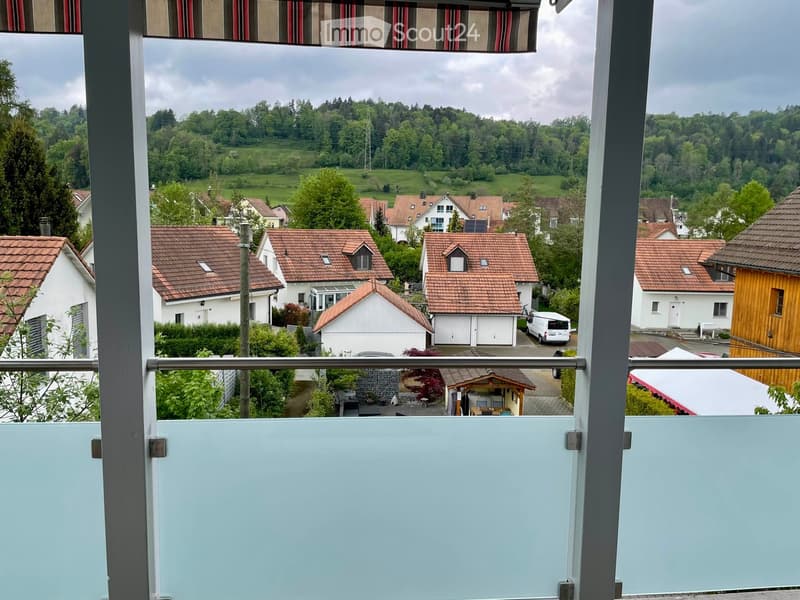Balcony overlooking a residential area with houses, trees, and a distant green landscape.