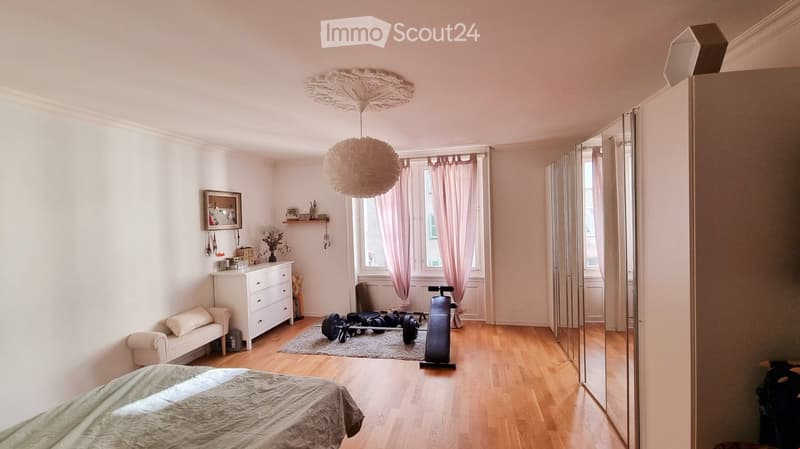 Well-lit bedroom with wooden flooring, a bed, a small sofa, a dresser, a mirror, and exercise equipment including a bike and weights.