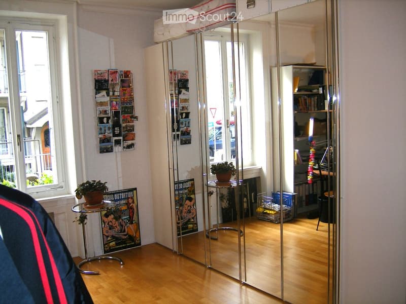 A modern, well-lit room with wooden flooring, large mirrors, a small round table with a potted plant, posters on the wall, and a desk area with shelves.