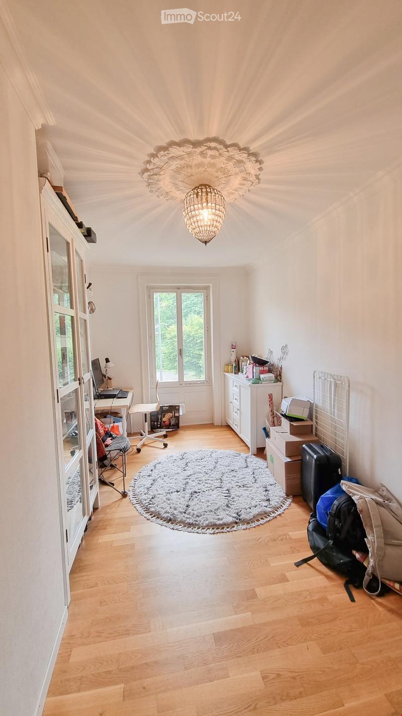 Bright room with wooden flooring, large window, chandelier, desk with laptop, chair, cabinet, and luggage near the wall.