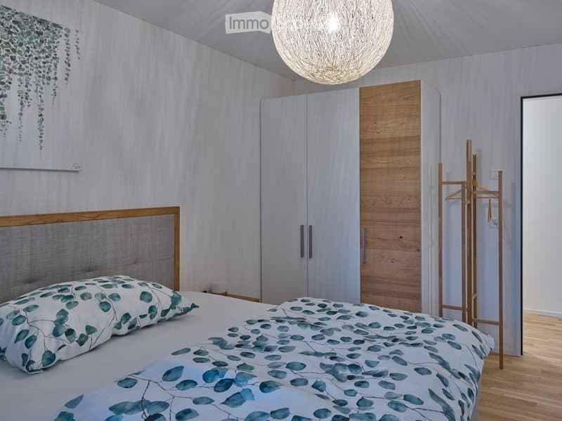 Modern bedroom with a wooden bed frame, leafy patterned bedding, white wardrobe, and a large spherical pendant light.