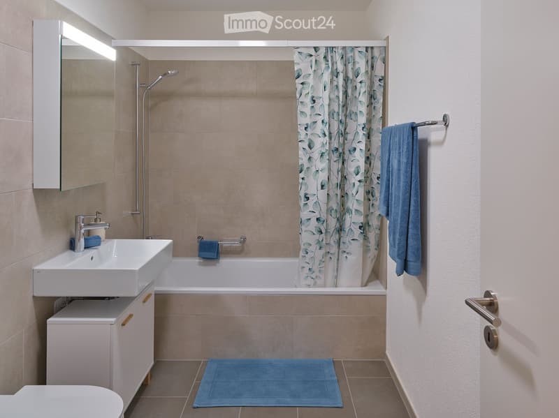 Modern bathroom with a white sink, bathtub, mirror, and blue towels. Features a leaf-patterned shower curtain and a blue bath mat.