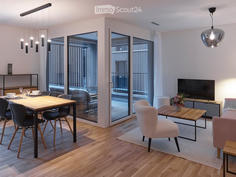 Modern open-plan living and dining area with wooden flooring, large windows, and a balcony view. Features a sofa, coffee table, dining table with chairs, and a TV stand.
