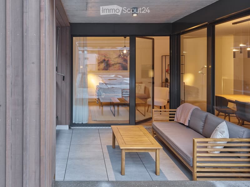 Modern balcony with wooden furniture, including a sofa, coffee table, and chairs, with large glass doors leading to a well-lit interior space.