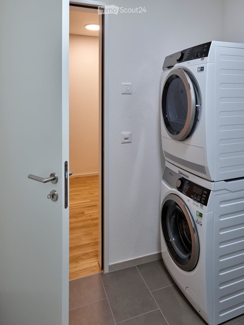 Modern bathroom with a stacked washer and dryer unit, grey tiled floor, white walls, and a wooden floor visible through an open door.