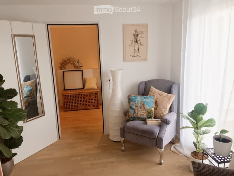 Cozy living room with a grey armchair, decorative pillows, a tall white vase, a large mirror, a small table with plants, and a framed skeleton print on the wall.