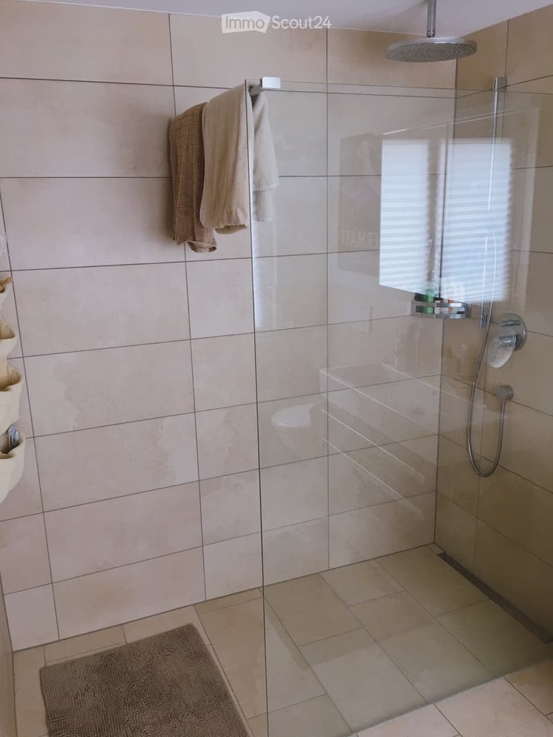 Modern bathroom with beige tiles, glass shower enclosure, towel rack, and a small shelf holding toiletries.
