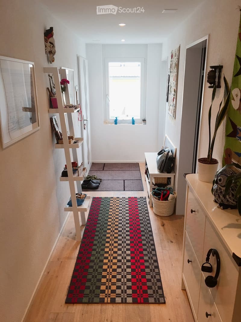 Bright hallway with wooden flooring, colorful checkered runner, shelves with decorative items, and a small table with bags and a potted plant.