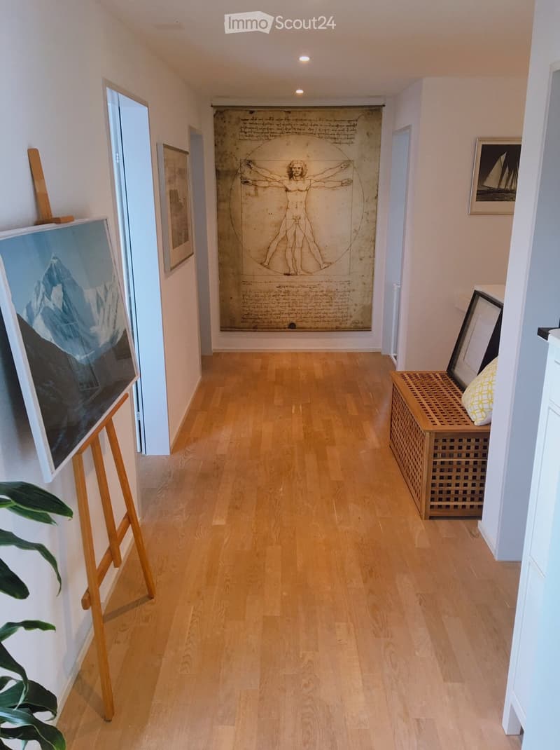 A narrow hallway with light wooden flooring, white walls, and several framed artworks and a large drawing on the wall. A small wooden bench with a cushion is placed near the end of the hallway.