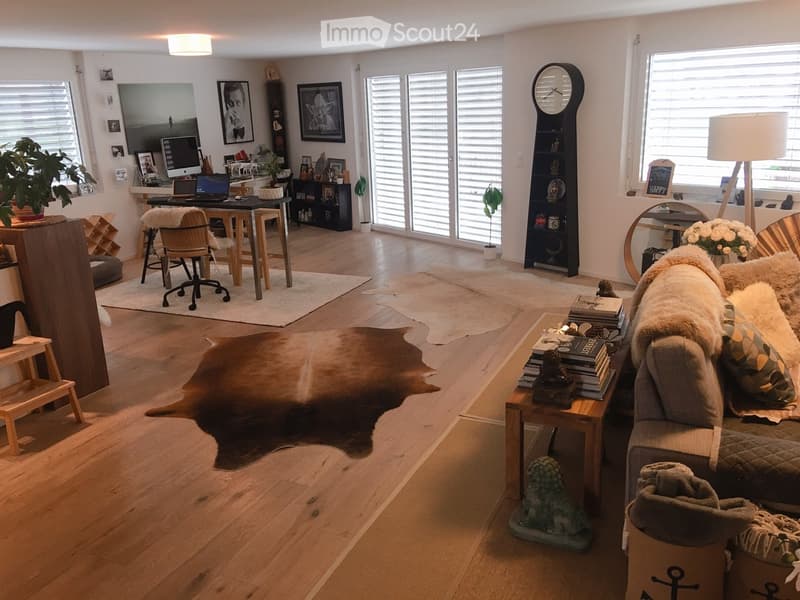 Living room with hardwood floors, large windows, a sofa with fur throw, coffee table, desk area with laptop, and decorative items including a cowhide rug and books.
