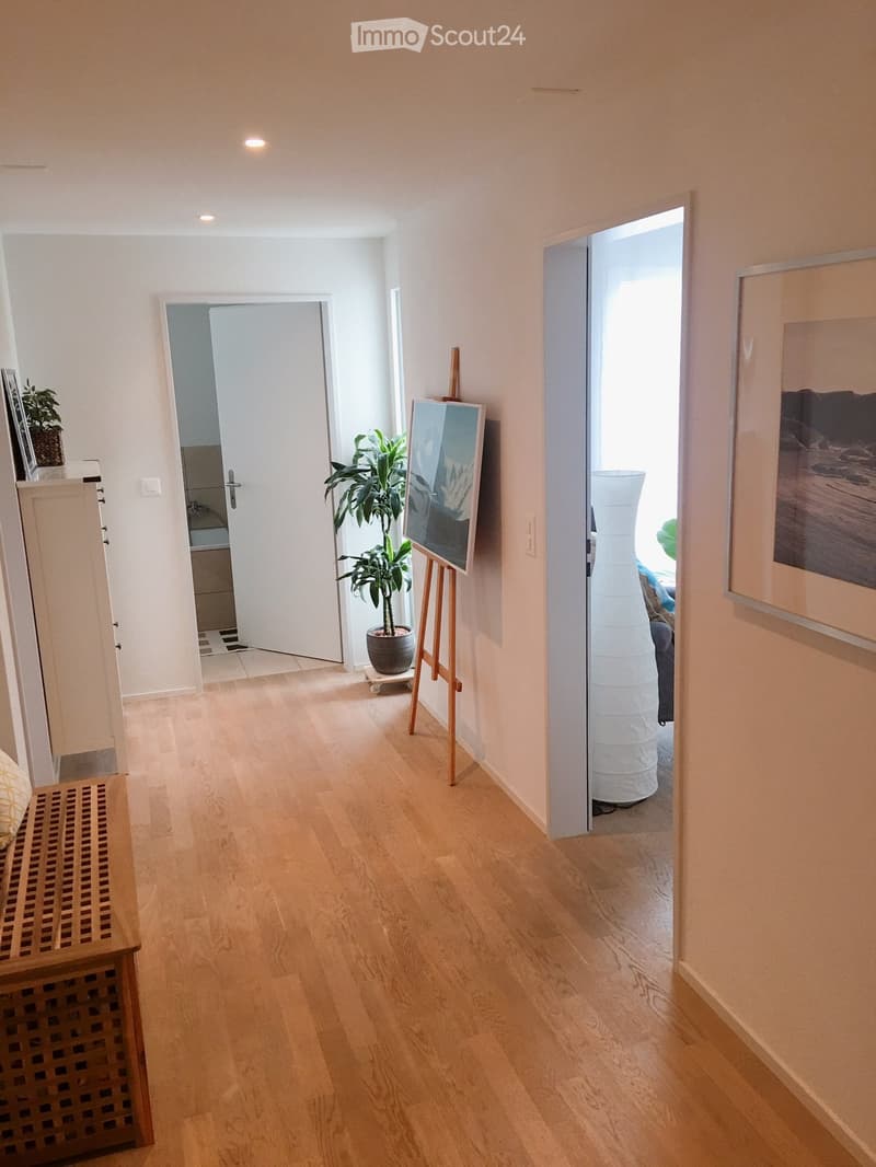 Bright hallway with light wooden flooring, white walls, and recessed lighting. Features include a small plant on a stand, an easel with a canvas, a tall white vase, and a partially visible bench on the left side.