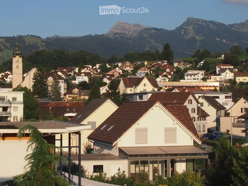 A panoramic view of a town with numerous houses, a church tower, and surrounding mountains under a clear blue sky.