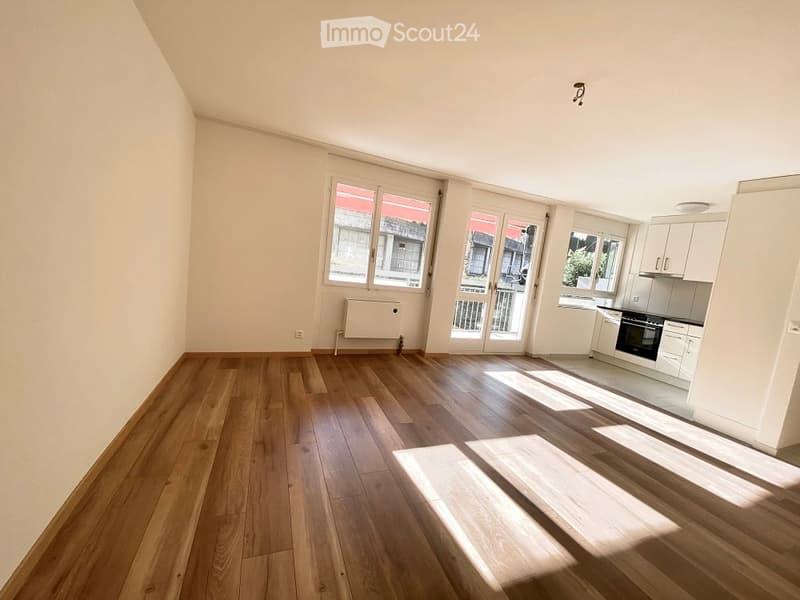 Bright, open-plan living and dining area with wooden flooring, large windows, and a small kitchen area featuring white cabinets and an oven. The space includes a balcony accessible through glass doors.