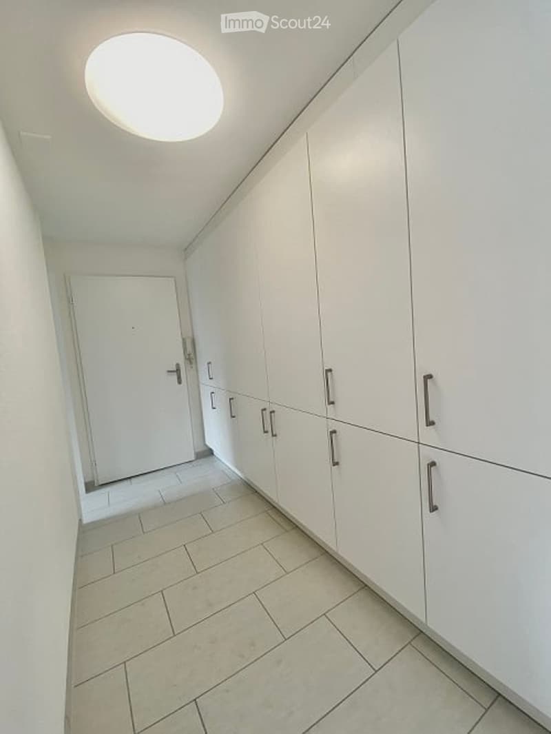 A clean, modern hallway with white tiled flooring, white walls, and built-in white cabinetry with silver handles. A closed white door is visible at the end of the hallway, and a round skylight provides natural light.
