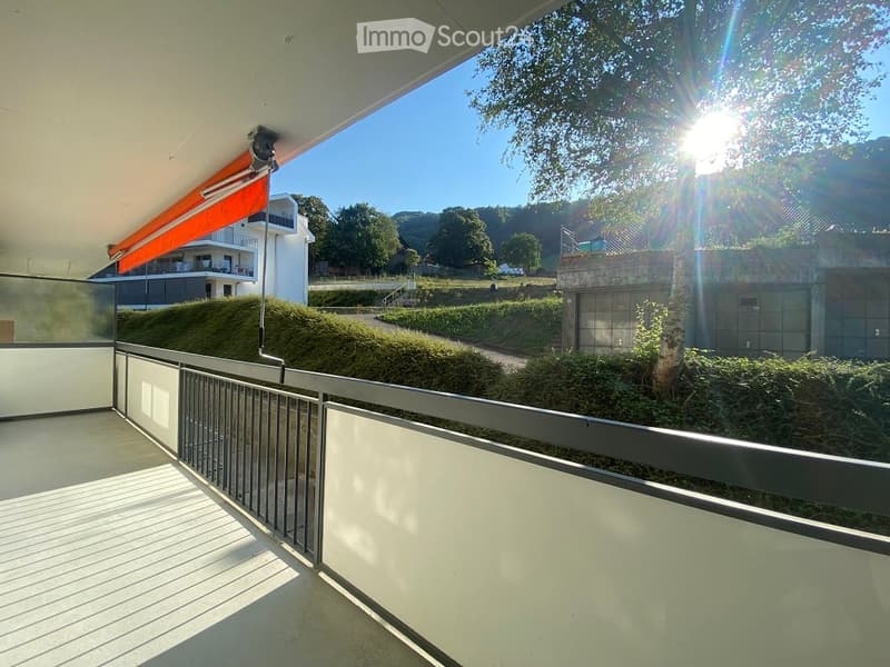 Balcony with metal railing, overlooking a landscaped area with trees and a modern building in the background.