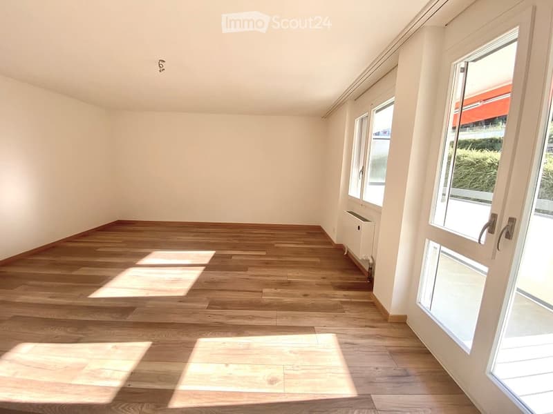 Empty room with wooden flooring and large windows allowing natural light to enter. The walls are painted white, and there are no furnishings or decorations visible.