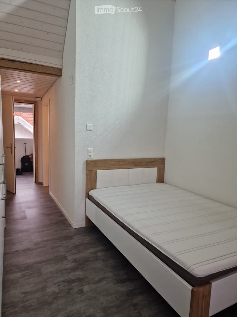Cozy bedroom with a single bed, wooden headboard, and white bedding. The room features light-colored walls, wooden flooring, and a small hallway leading to another area.