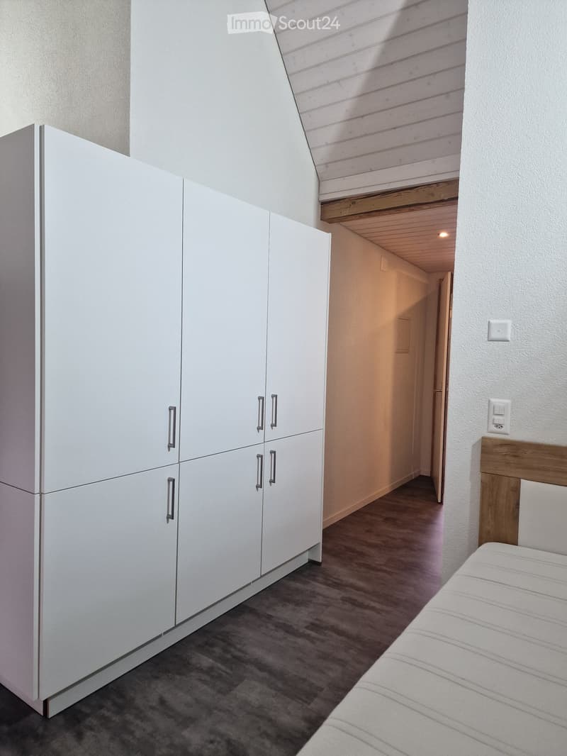 Modern bedroom with a white wardrobe, a neatly made bed, and a wooden slanted ceiling. The room features a dark grey carpet and white walls.