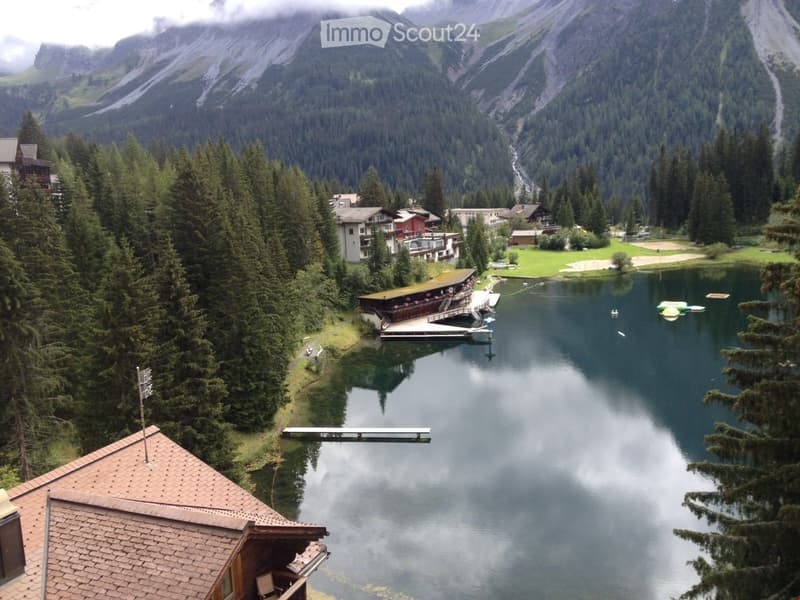 A scenic view of a lake surrounded by dense forest and mountains, with a dock extending into the water and several buildings nearby.