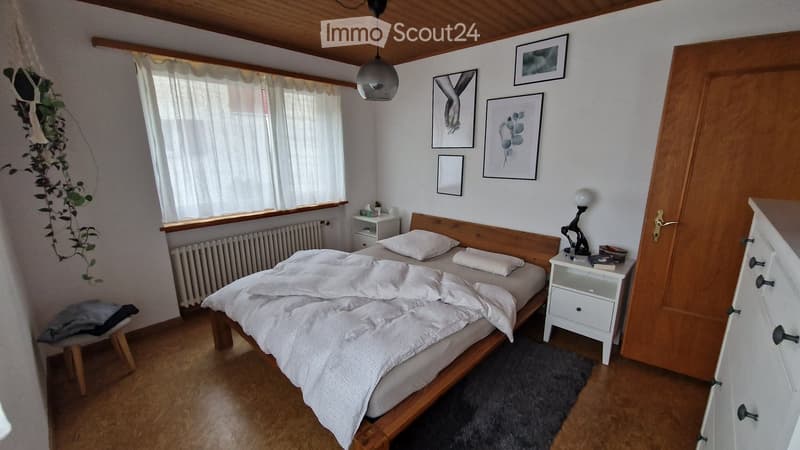 Bedroom with bed, nightstands, radiator, framed pictures, wooden floor, window with curtains, plant, stool, wooden door, dresser