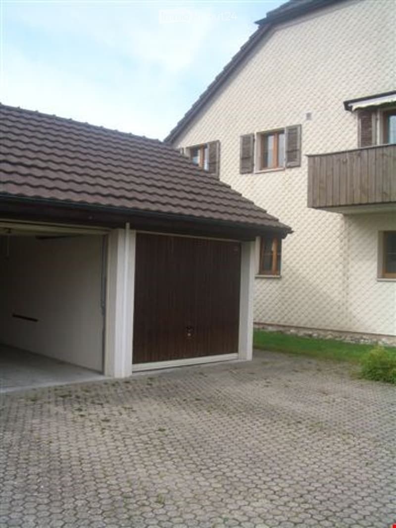 Two-story residential building with a garage and balcony, featuring a tiled roof and light-colored facade.