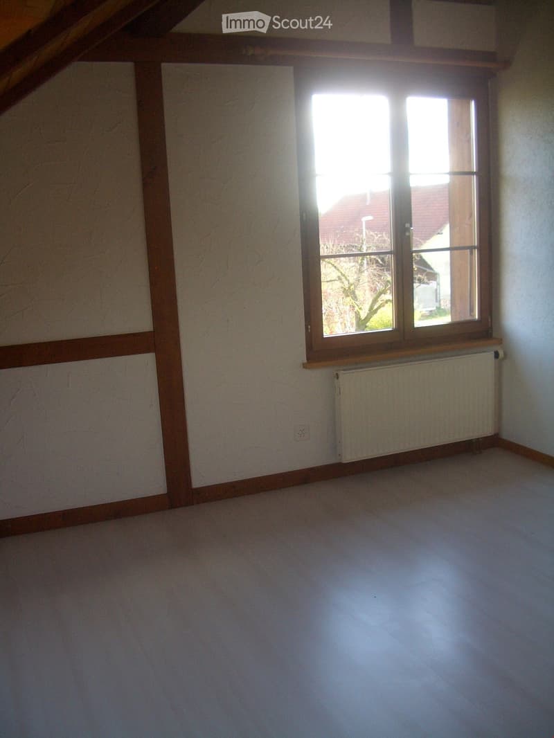 Empty attic room with wooden beams, white walls, tiled floor, and a large window with wooden frame and radiator below it.