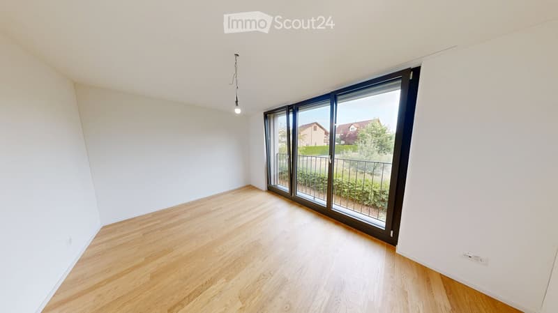 Empty room with light wooden flooring, white walls, and large sliding glass doors leading to a balcony with a view of greenery and neighboring houses.