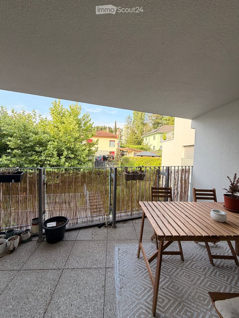 Balcony with wooden table, chairs, and potted plants, overlooking a green area with trees and houses.