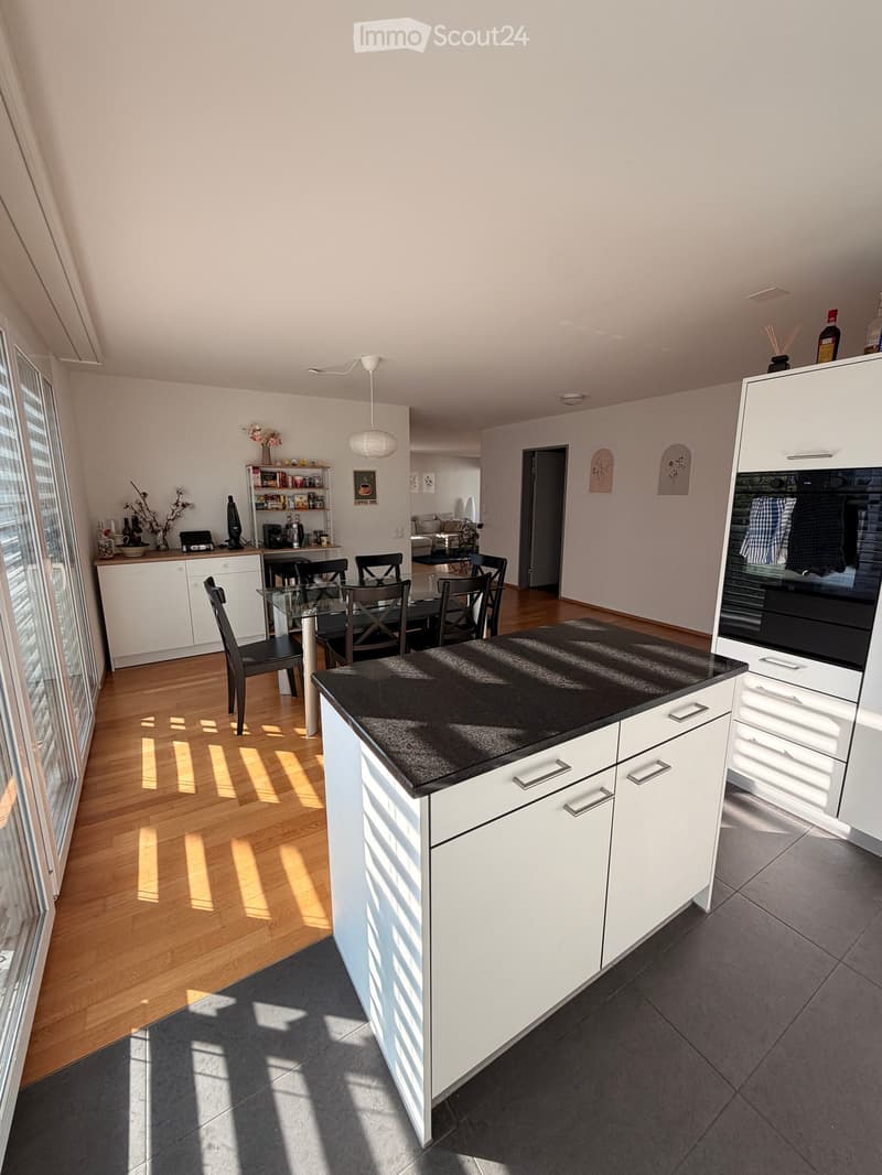 Modern kitchen with white cabinets, black countertop, glass dining table, and chairs. Features a built-in oven and a small living area visible in the background.
